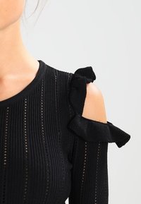 Zwarte gebreide top met verticale ribbels, voorzien van een cold shoulder ontwerp en gerimpelde accenten bij de mouwen. Gelaagd stof met geperforeerde details.