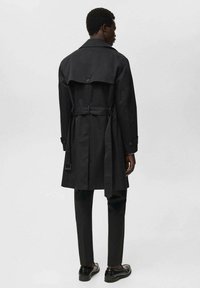 Abrigo negro de trench con detalle de botón en la parte trasera, cintura ceñida con cinturón y una ligera abertura. Tejido mate, puños abotonados, combinado con pantalones y zapatos negros.