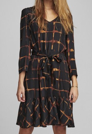 Kobieta ubrana w czarną sukienkę z wzorem kratki w kolorze rdzawego pomarańczu, z dekoltem w serek, długimi rękawami, paskiem zawiązanym w talii oraz falbaną u dołu, stojąca na jednolitym tle.