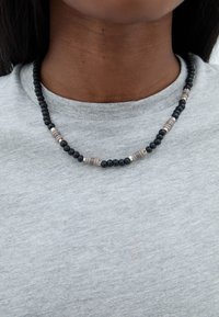 Fekete gyöngyös nyaklánc váltakozó fém és textúrált gyöngyökkel, szürke szöveten viselve. Egyszerű kialakítás, eltérő gyöngymérettel és formákkal.