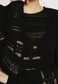 Femme portant un haut en dentelle noire à manches longues avec des motifs géométriques et floraux, partiellement transparent, main reposant près de son visage.