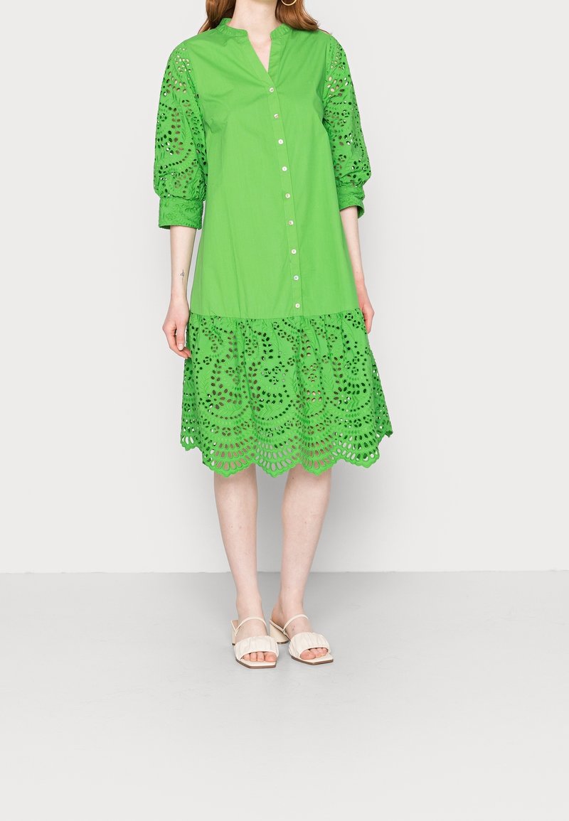 Mujer vestida con un vestido botoné brillante de color verde con patrones de encaje en las mangas y el dobladillo, combinado con sandalias de tacón de bloque abiertas en la punta de color blanco.