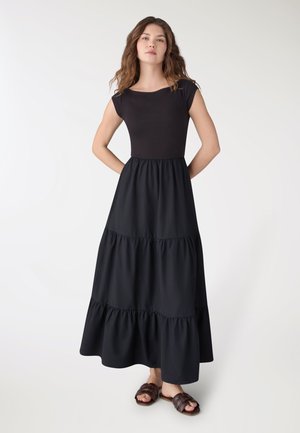 Kobieta stojąca z rękami za plecami, ubrana w czarną, warstwową sukienkę maxi z krótkimi rękawami typu czapka i brązowe plecione sandały na płaskim obcasie na białym tle.