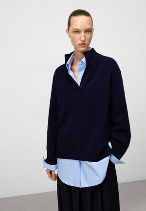 Maglione lavorato a maglia blu scuro con collo alto sopra una camicia azzurro chiaro con bottoni. Il maglione presenta polsini a coste e una silhouette oversize.
