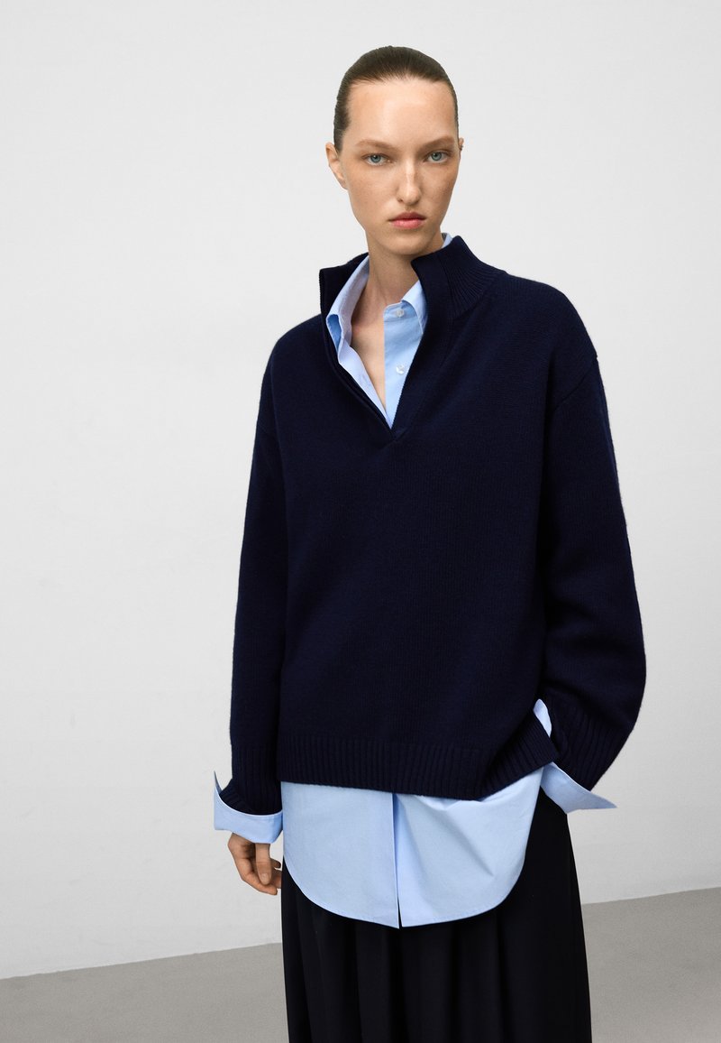 Maglione lavorato a maglia blu scuro con collo alto sopra una camicia azzurro chiaro con bottoni. Il maglione presenta polsini a coste e una silhouette oversize.