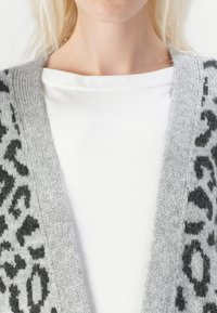 Cardigan grigio con bordi a stampa leopardata nera, texture lavorata a maglia e apertura ampia davanti. Indossato su una camicia bianca semplice.
