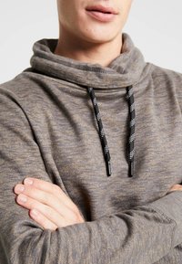 Grå hættetrøje med tragtkrave, tekstureret stof med guldpletter. Sorte snore med hvide accenter. Modellen har armene tværs.