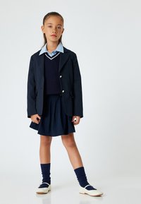 Blazer marine avec jupe assortie,superposé à une chemise bleu clair et un pull marine. Des chaussures blanches et des chaussettes bleues complètent la tenue.