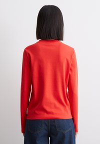 Rød langærmet skjorte lavet af bomuld, med rund hals og en glat tekstur. Skjorten er kombineret med blå jeans.