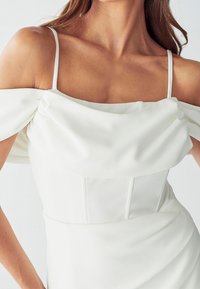 Donna che indossa un vestito bianco off-shoulder con sottili spaghettini e dettaglio in tessuto pieghettato attorno al petto e alle maniche.