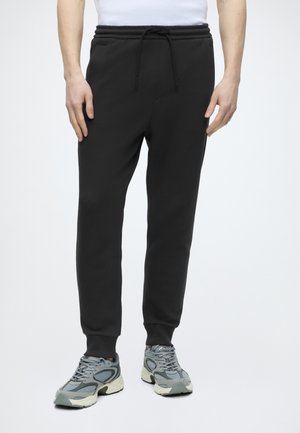 Homme portant un pantalon de jogging noir à taille coulissée et des baskets de sport grises, debout devant un fond blanc uni.