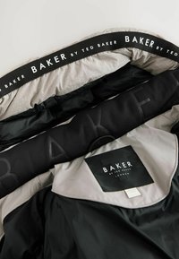 Sort og grå dunjakke indvendigt med en quiltet tekstur, der har et fremtrædende "BAKER by Ted Baker" mærkat og iøjnefaldende sorte accenter.