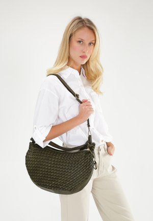 Olijfgroene leren handtas van gevlochten leer met een gewelfde vorm, een verstelbare schouderband en decoratieve hardwareaccenten. Model draagt een witte blouse en broek.