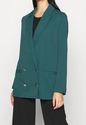 Vrouw draagt een teal double-breasted blazer met zilveren knopen over een zwart overhemd en zwarte broek, afgebeeld van nek tot midden van het bovenbeen.