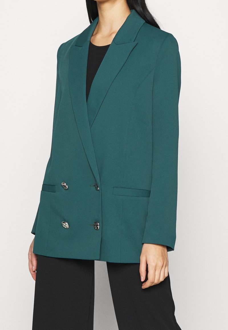 Femme portant un blazer croisé couleur sarcelle avec des boutons argentés sur une chemise noire et un pantalon noir, montrée du cou jusqu'à mi-cuisse.