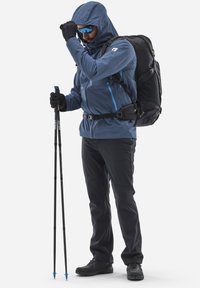 Veste bleue avec capuche, pantalon noir et sac à dos noir. Gants et lunettes de soleil portés. Utilisation de bâtons de trekking avec des pointes bleues.