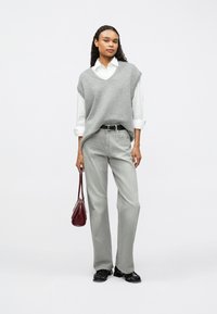 Γυναίκα ντυμένη με άσπρο πουκάμισο με διπλωμένα μανίκια, γκρίζο αμάνικο πουλόβερ, ανοιχτό γκρι τζιν, μαύρα παπούτσια, και κρατώντας μια μικρή μπορντώ τσάντα.