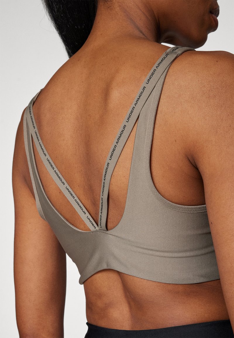 Espalda superior de una mujer que lleva un sujetador deportivo color topo con doble tirante, un juego liso y el otro impreso con el texto "Under Armour".