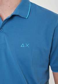 Polo shirt blu realizzato in tessuto testurizzato, con colletto turchese a contrasto e un piccolo ricamo sul lato sinistro del petto.