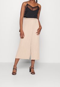 Svart cami-top med spetsdetaljer som kombineras med beige plisserade culotter. Modellen bär svarta klacksandaler med blockhäng. Enkelt, flödande mönster och lätt textur.