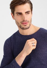 Homem a usar uma camisa de manga longa azul marinho texturizada, segurando uma aliança metálica de múltiplas bandas com a mão esquerda, pele suave e cabelo curto.