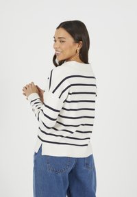 Kobieta z ciemnymi włosami ubrana w biały sweter w czarne poziome paski i niebieskie dżinsy, uśmiechająca się i patrząca w bok na tle białym.