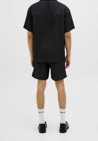 Uomo vestito con una maglietta nera a coste a maniche corte e pantaloncini abbinati, calze bianche con marchio e scarpe nere, in piedi contro uno sfondo bianco uniforme.