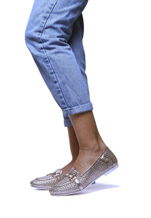 Mocassini intrecciati in oro metallico con tacco basso, caratterizzati da una fibbia decorativa e fianchi lisci. Indossati con pantaloni in denim blu arrotolati.