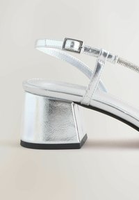 Silberne Sandalen mit Absätzen verfügen über eine glänzende metallische Textur, einen quadratischen Blockabsatz und verstellbare Knöchelriemen mit einer Schnallenverschluss.