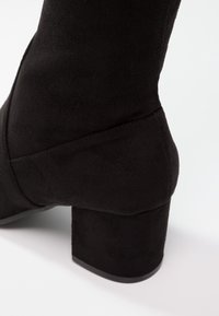 Bottine en suède noir avec un talon large et carré. Présente une surface lisse et texturée ainsi qu'un design élégant et effilé à la cheville.