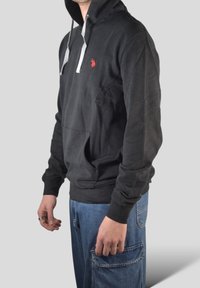 Czarna bawełniana bluza z kapturem z szarymi sznurkami, przednią kieszenią kangurką i małym czerwonym logo na piersi; zestawiona z niebieskimi dżinsowymi spodniami typu cargo.