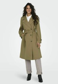 Olivgrön trenchcoat med dubbelknäppta knappar, ett bälte och sidofickor, tillsammans med ljusgrå uppvikta byxor och svarta ankelskärp.