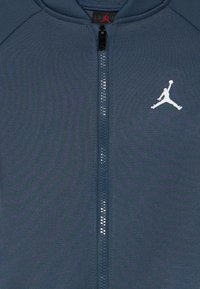 Μπλε μπουφάν με πλήρη φερμουάρ και λευκό λογότυπο Jumpman στο αριστερό στήθος και μαύρη λαβή φερμουάρ με κόκκινη σιλουέτα Jumpman.