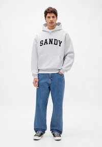 Szara bluza z kapturem z napisem "SANDY" w ciemnym kolorze, przednia kieszeń, w połączeniu z luźnymi niebieskimi dżinsami i czarno-białymi sneakersami w wzory.