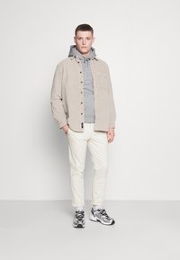 Beige kordfløjl skjorte, grå hættetrøje, cremefarvede bukser og grå-sorte sneakers. Skjorten har en lomme foran og knaplukning.