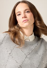 Grauer Pullover mit einem Rautenmuster aus Pailletten, mit einem gerippten Kragen und darüber getragen über einem weißen Hemd mit Kragen. Weiche Textur.