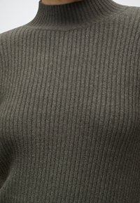 Gros plan d'une personne portant un pull à col roulé côtelé vert olive avec une texture fine en tricot.