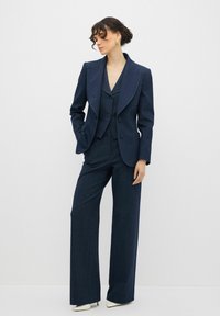 Mörkblå skräddarsydd kostym med en strukturerad kavaj, en ärmlös väst och vida byxor, tillverkad av textil med struktur; accentuerad med vita klackar.