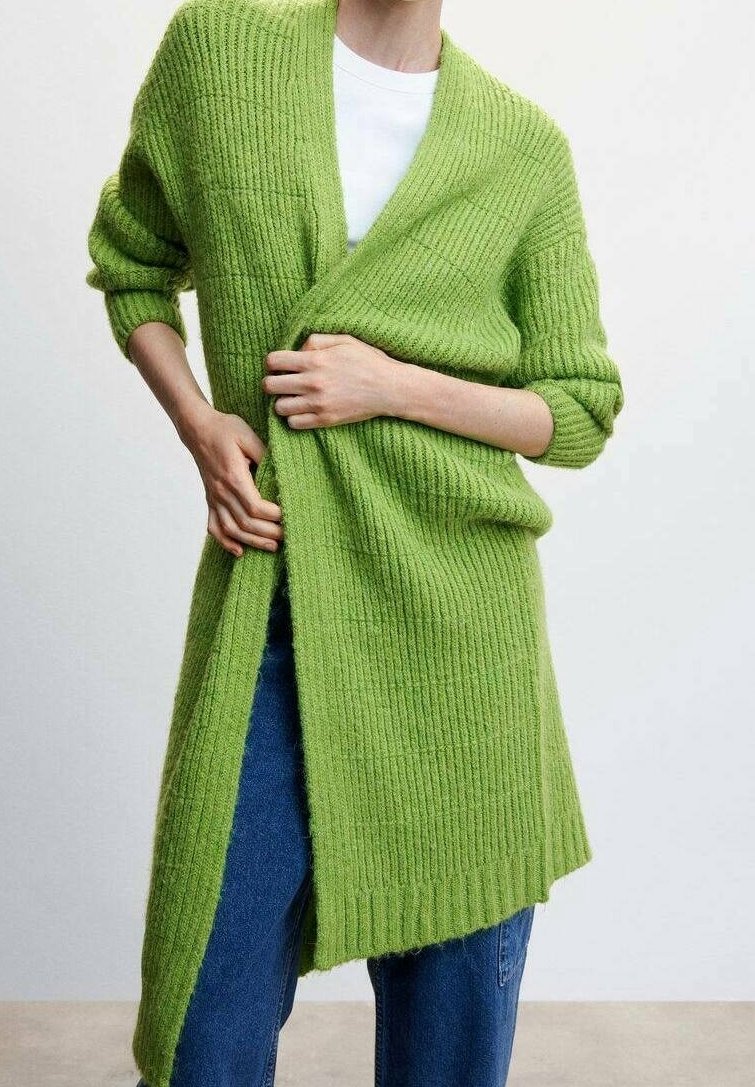 Personne portant un long cardigan vert côtelé sur une chemise blanche et un jean bleu, ajustant le cardigan avec les deux mains.