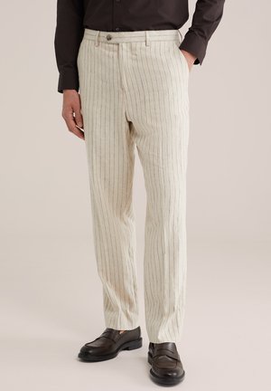 Uomo che indossa pantaloni beige a righe sottili, camicia marrone scuro con bottoni e mocassini in pelle marrone scuro, in piedi con una mano in tasca.