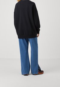 Persona che indossa un top nero oversize a maniche lunghe, jeans a gamba larga blu e sneakers nere e arancioni, in piedi contro uno sfondo semplice.