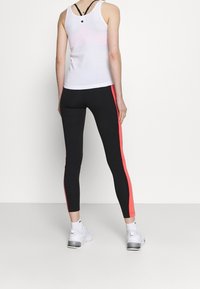 Person i svarta leggings med rosa sidostreck, en vit linnetröja med en logotyp på övre delen av ryggen, och vita sneakers, står vänd bort.