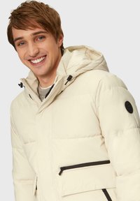 Puffermantel in Creme mit abnehmbarer Kapuze, ausgestattet mit schwarzen Reißverschlüssen und einem Logopatch am Ärmel. Glatte Textur, gestepptes Design.