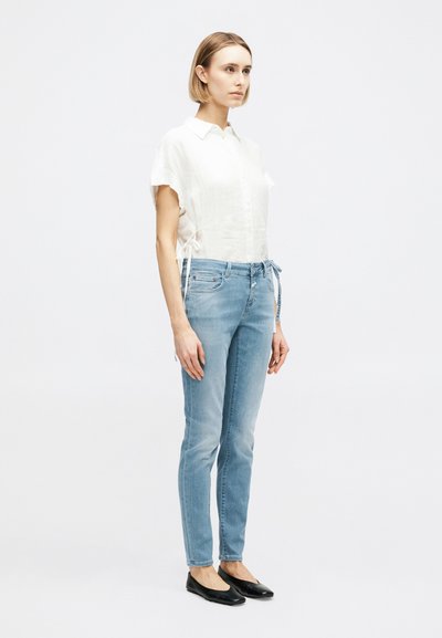 Femme debout portant une chemise blanche à manches courtes avec boutons, un jean bleu clair et des chaussures plates noires, sur un fond clair uni.