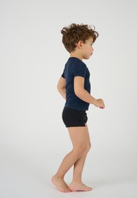 Camicetta blu navy a maniche corte abbinata a pantaloni neri, entrambi realizzati in tessuto morbido. Bambino di profilo, a piedi nudi, con capelli ricci.