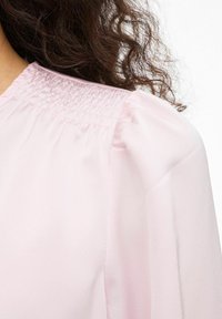 Blouse rose clair avec un col smocké, des manches bouffantes et un tissu lisse à la texture subtilement brillante.