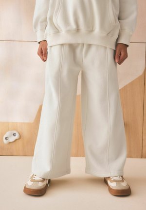 Person trägt cremefarbene weite Hosen, passenden oversized Sweatshirt und beige-weiße Sneaker und steht vor einer Holzwand.