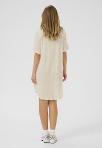 Robe beige à manches courtes avec un tissu léger et texturé, présentant un ourlet incurvé et des fentes latérales, associée à des baskets blanches.