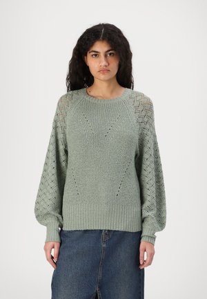 Pull en maille vert clair avec un ourlet côtelé, présentant un motif texturé et des détails en dentelle sur les manches. Assorti avec une jupe en denim.