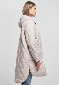 Quiltet beige jakke med hette, med en lang, buet kant og sidespalter. Laget av lett, polstret stoff.
